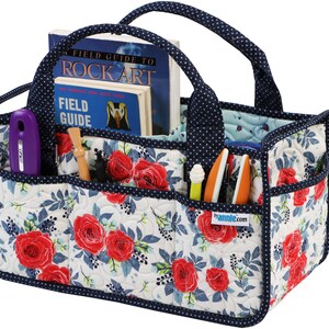 May include: A quilted storage tote with a navy blue handle and trim. The tote has multiple pockets and is decorated with a floral pattern of red roses and blue leaves on a white background. The tote is filled with various craft supplies, including a book titled "Field Guide to Rock Art", a purple tool, and several pens and pencils.
