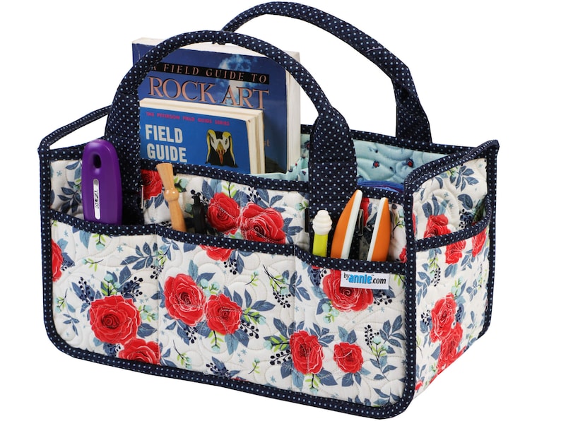 May include: A quilted storage tote with a navy blue handle and trim. The tote has multiple pockets and is decorated with a floral pattern of red roses and blue leaves on a white background. The tote is filled with various craft supplies, including a book titled "Field Guide to Rock Art", a purple tool, and several pens and pencils.