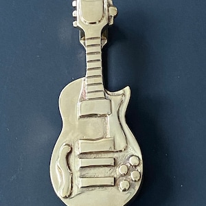 May include: A gold-colored metal door knocker shaped like an electric guitar. The guitar has a detailed design with a pickguard, knobs, and a fretboard.