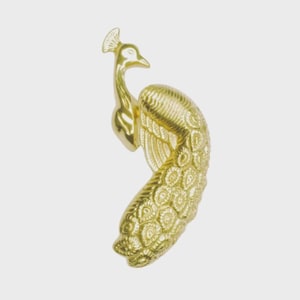May include: A gold-colored peacock figurine with detailed feather patterns. The bird is depicted in a curved pose, with its head and tail feathers prominently displayed. The figurine has a smooth, polished finish and is set against a plain background.