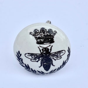 以下が含まれることがあります： 白いセラミック製のノブで、黒と白のデザインで、王冠をかぶった蜂が描かれています。蜂は葉の輪で囲まれています。
