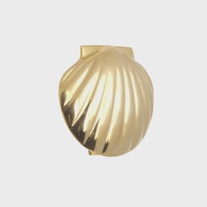 May include: A gold-colored, seashell-shaped decorative object. The shell has a ridged texture and a small, rectangular attachment at the top. The object is likely made of metal and has a reflective surface.