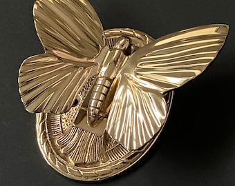 May include: A gold-colored metal butterfly-shaped decorative object. The butterfly has detailed wing patterns and is mounted on an oval base with a textured border. The object is set against a dark background.
