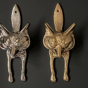 May include: Two metal fox-shaped wall hooks. One is silver-toned, the other is gold-toned. Each hook features a detailed fox head with a decorative feather-like element above. The hooks have a loop at the top for hanging.