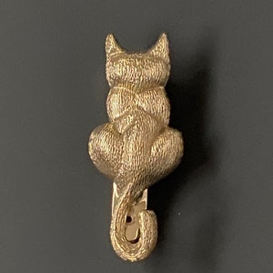 May include: A gold-colored cat-shaped hair clip. The clip is in the shape of a cat sitting with its back to the viewer. The cat's tail curls down and around to form the clip's clasp.