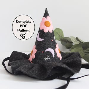 May include: A dark gray felt witch hat with a wide brim and a pointed top. The hat is decorated with pink flowers, white crescent moons, and a white star. The text "Complete PDF Pattern" is in a circle on the left.