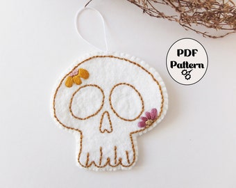 May include: White felt skull ornament with embroidered details. The skull has two yellow flowers and one pink flower on it. The ornament has a white string loop for hanging.