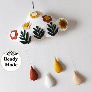 May include: A white felt cloud mobile with felt flowers in yellow, orange, and red, and green leaves.  The cloud has four felt raindrops hanging from it in yellow, white, orange, and brown.