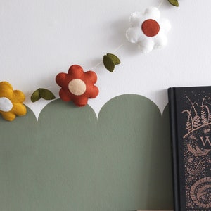 May include: A felt flower garland with yellow, white, and red flowers, and green leaves. The garland is strung against a white wall with a green scalloped border. A book with gold lettering sits on a wooden shelf.