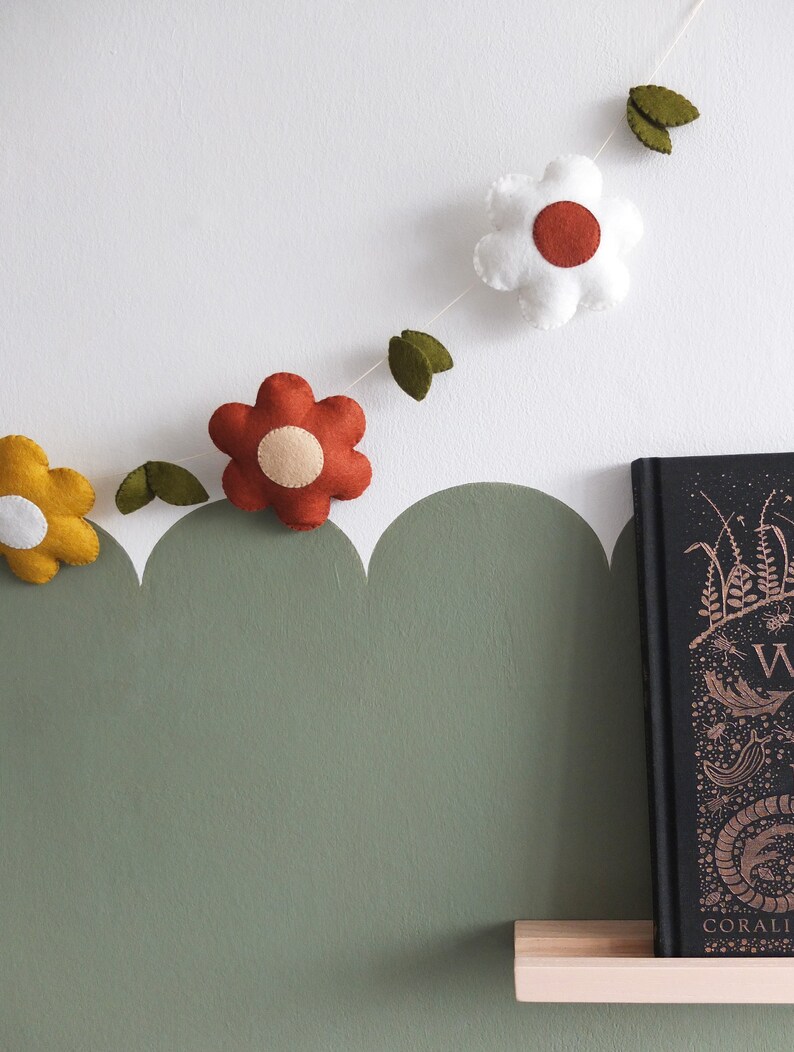 May include: A felt flower garland with yellow, white, and red flowers, and green leaves. The garland is strung against a white wall with a green scalloped border. A book with gold lettering sits on a wooden shelf.