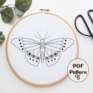 以下が含まれることがあります： 木製の刺繍枠の中に白い布地に描かれた蝶の白黒線画。画像の右下隅に「PDF Pattern」の文字があります。