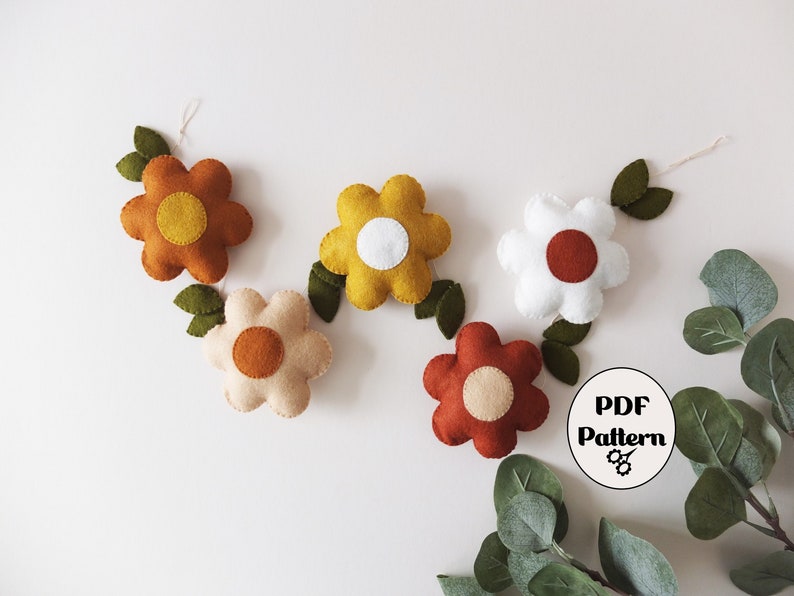 May include: A felt flower garland featuring five colorful flowers in shades of orange, yellow, white, and red-brown, each with a contrasting center. Green felt leaves and a PDF pattern icon are also visible.