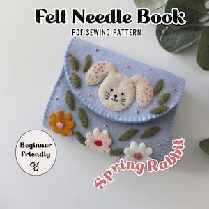 May include: A light blue felt needle book with a bunny design and floral accents. The book is labeled "Felt Needle Book" and "PDF Sewing Pattern". The design includes a bunny face, flowers, and the text "Spring Rabbit" and "Beginner Friendly".