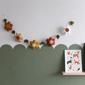 May include: A felt flower garland with various colored flowers, including orange, yellow, and white, hangs against a white wall. Green felt leaves are interspersed between the flowers. A framed print of woodland animals sits on a shelf below.