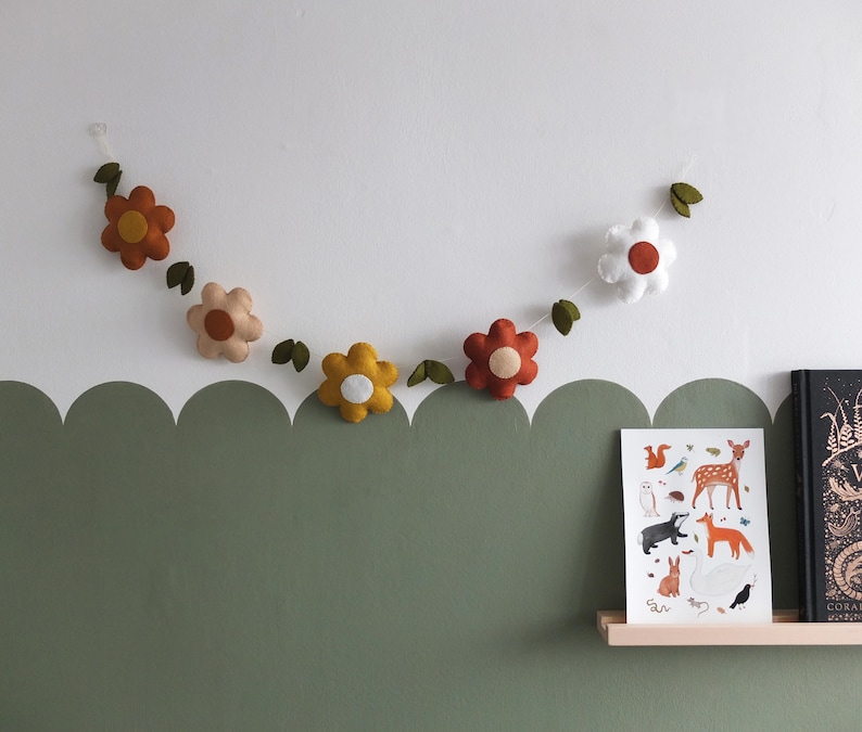 May include: A felt flower garland with various colored flowers, including orange, yellow, and white, hangs against a white wall. Green felt leaves are interspersed between the flowers. A framed print of woodland animals sits on a shelf below.