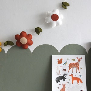 May include: A felt flower garland with white and rust-colored flowers and green leaves hangs above a framed woodland animal print. The print features illustrations of a deer, fox, owl, and other forest creatures. The wall is painted white and olive green.