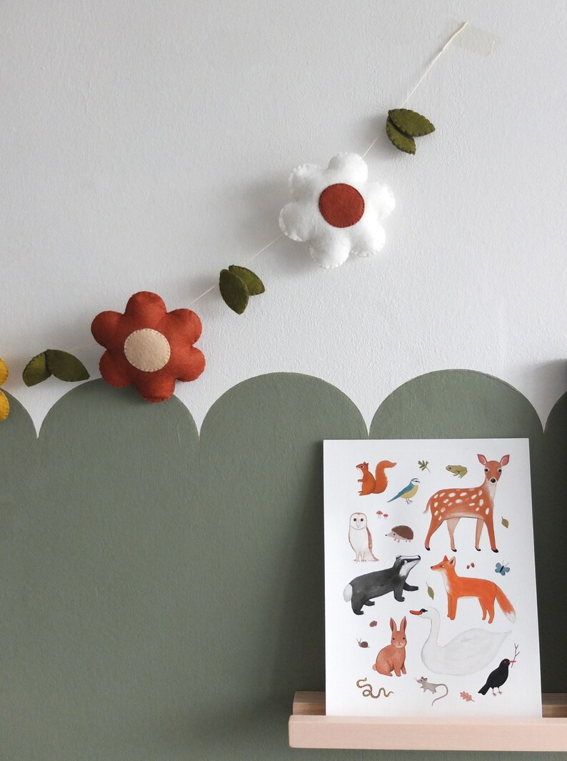 May include: A felt flower garland with white and rust-colored flowers and green leaves hangs above a framed woodland animal print. The print features illustrations of a deer, fox, owl, and other forest creatures. The wall is painted white and olive green.