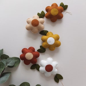 May include: A garland of felt flowers in various colors, including white, beige, mustard yellow, and rust. Each flower has a contrasting center and is connected by green felt leaves. The flowers are arranged on a white background.