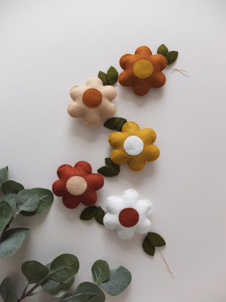May include: A garland of felt flowers in various colors, including white, beige, mustard yellow, and rust. Each flower has a contrasting center and is connected by green felt leaves. The flowers are arranged on a white background.