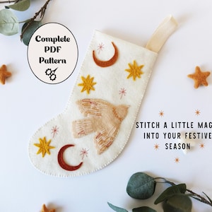 Könnte beinhalten: Ein weißer Weihnachtsstrumpf aus Filz mit gestickten Himmelsmotiven, darunter ein Vogel, Halbmonde und Sterne. Der Text "Complete PDF Pattern" befindet sich in einem Kreis. Der Satz "Stitch a little magic into your festive season" ist ebenfalls vorhanden.
