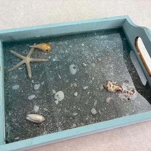Op de afbeelding: Een rechthoekig lichtblauw houten dienblad met uitgesneden handgrepen. De bodem is voorzien van een heldere harsinleg met grijs zand, een lichtbruine zeester en diverse kleine witte en bruine schelpen verspreid over het oppervlak.