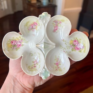 May include: A white porcelain serving dish with a clover-like design. Each of the four sections features a floral pattern with pink roses and green leaves. The dish is held in a hand, showcasing its delicate details.
