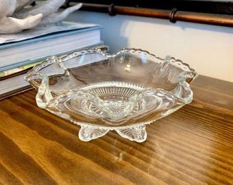 May include: A clear glass footed bowl with a scalloped edge and intricate cut glass design. The bowl is sitting on a wooden surface.