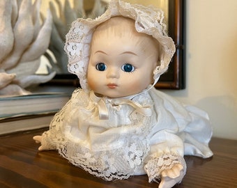 Könnte beinhalten: Eine Porzellan-Babypuppe, die ein weißes Spitzenkleid und eine Haube trägt. Die Puppe hat blaue Augen und ein gemaltes Lächeln.