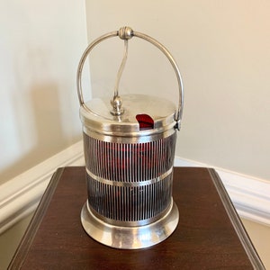 May include: Silver sugar dispenser with a red glass insert and a wire mesh design. The dispenser has a handle and a lid.