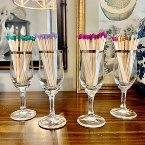 May include: Five clear glass stemmed shot glasses filled with colorful matchsticks. The matchsticks have colored tips in shades of purple, teal, pink, and orange.
