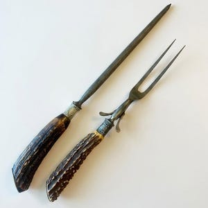 May include: An antique carving set featuring a sharpening steel and a carving fork. The handles are made of dark brown, textured material, possibly antler, with ornate silver-colored metal accents. The steel and fork are a dark, aged metal.