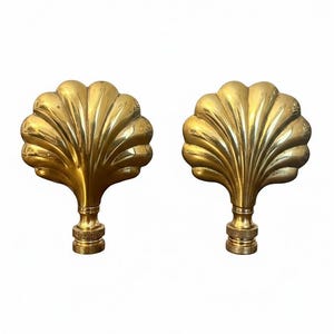 May include: Two brass-colored lamp finials with a seashell design. Each finial has a detailed, fan-shaped top and a threaded base for attachment. The finials are displayed against a white background.