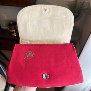K&ouml;nnte beinhalten: Eine rote Clutch aus Satin mit einem goldenen Stickmuster aus zwei Federn. Die Clutch hat ein wei&szlig;es Satin-Futter und einen silbernen Druckknopfverschluss.