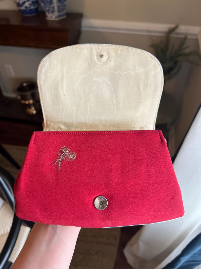 K&ouml;nnte beinhalten: Eine rote Clutch aus Satin mit einem goldenen Stickmuster aus zwei Federn. Die Clutch hat ein wei&szlig;es Satin-Futter und einen silbernen Druckknopfverschluss.