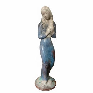 May include: A ceramic figurine of a woman in a long blue dress, with her hands clasped in front of her. The figure has long, flowing hair and stands on a round base. The dress has a marbled effect.