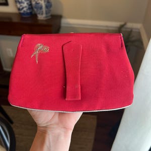 K&ouml;nnte beinhalten: Eine rote Stoff-Clutch mit einem goldenen und wei&szlig;en Pfauenfeder-Design. Die Clutch hat einen Klappverschluss mit einer kleinen rechteckigen Schnalle.