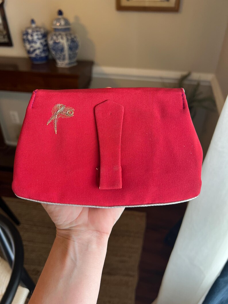 K&ouml;nnte beinhalten: Eine rote Stoff-Clutch mit einem goldenen und wei&szlig;en Pfauenfeder-Design. Die Clutch hat einen Klappverschluss mit einer kleinen rechteckigen Schnalle.