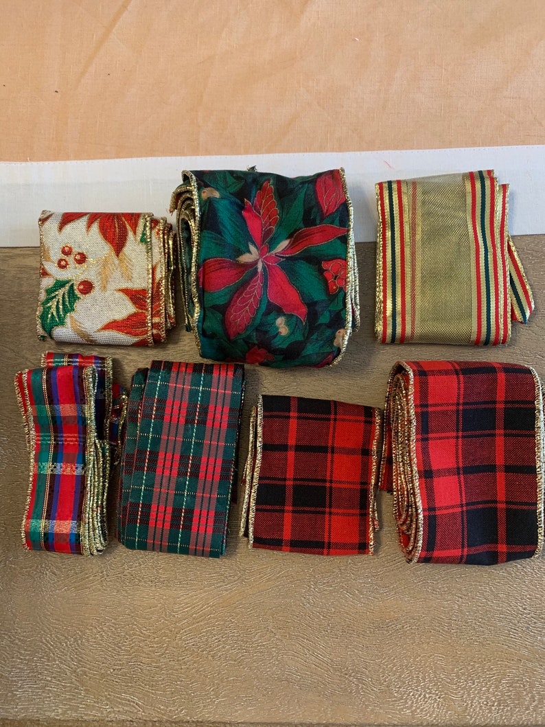 May include: Assortment of seven different Christmas ribbon rolls. The ribbons are red, green, gold, and white with various patterns including plaid, floral, and stripes. The ribbons have gold trim.