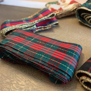 May include: A close-up of a red and green plaid ribbon with gold trim. The ribbon is folded and stacked on a wooden surface.