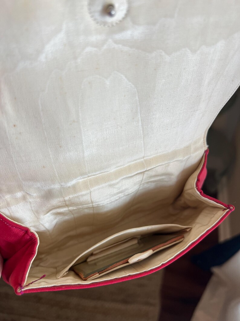 K&ouml;nnte beinhalten: Eine rote Clutch aus Satin mit einem wei&szlig;en Satin-Innenfutter. Die Clutch hat eine kleine Tasche im Inneren.