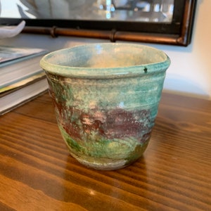 May include: A small, green, brown, and white ceramic cup with a glazed finish. The cup is sitting on a wooden surface.