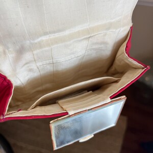 K&ouml;nnte beinhalten: Eine rote Samt-Abendtasche mit einem kleinen, rechteckigen Spiegel, der an der Vorderseite befestigt ist. Die Tasche ist ge&ouml;ffnet und zeigt ein wei&szlig;es Satin-Futter.