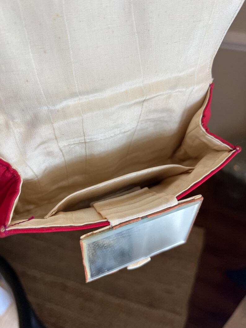 K&ouml;nnte beinhalten: Eine rote Samt-Abendtasche mit einem kleinen, rechteckigen Spiegel, der an der Vorderseite befestigt ist. Die Tasche ist ge&ouml;ffnet und zeigt ein wei&szlig;es Satin-Futter.