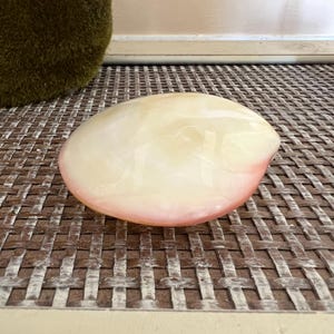 May include: A smooth, oval-shaped decorative object with a pearlescent finish. The object transitions from a creamy white to a soft pink hue along its edge. It rests on a woven, brown surface.