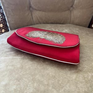 K&ouml;nnte beinhalten: Eine rote Clutch aus Satin mit einem silbernen Stickmuster. Die Clutch hat einen wei&szlig;en Rand.