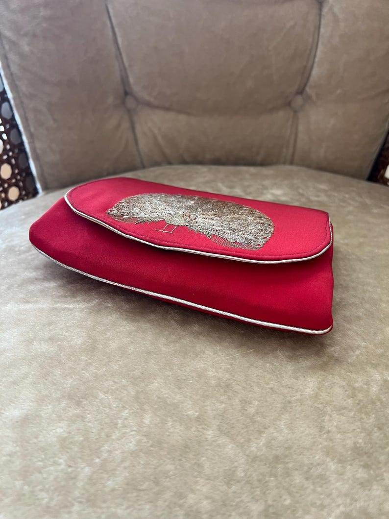 K&ouml;nnte beinhalten: Eine rote Clutch aus Satin mit einem silbernen Stickmuster. Die Clutch hat einen wei&szlig;en Rand.