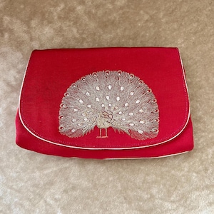 K&ouml;nnte beinhalten: Eine rote Clutch aus Satin mit wei&szlig;em Besatz und einem gestickten Pfauenmotiv in Goldfaden.