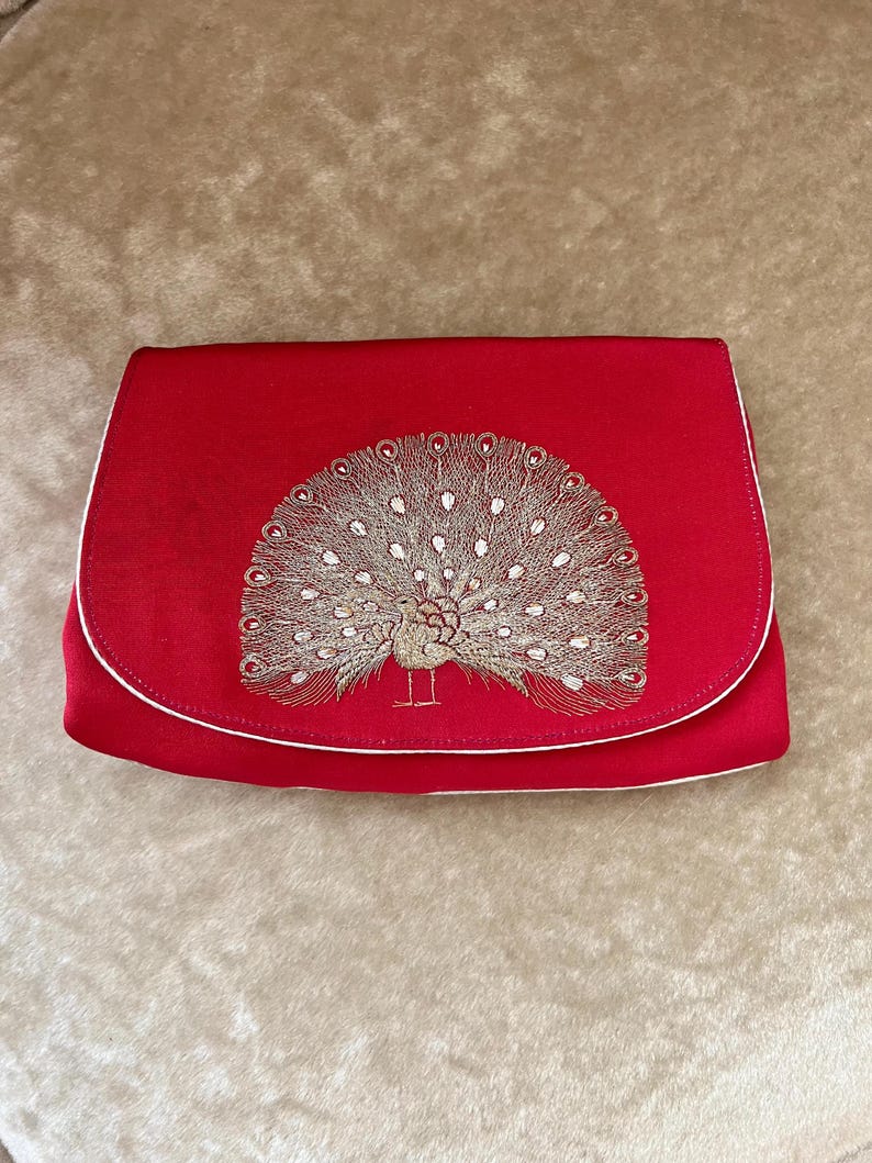 K&ouml;nnte beinhalten: Eine rote Clutch aus Satin mit wei&szlig;em Besatz und einem gestickten Pfauenmotiv in Goldfaden.