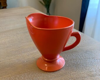 Könnte beinhalten: Ein leuchtend orangefarbener Keramik-Milchkännchen mit Henkel und einem Sockel.
