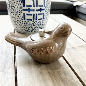 May include: A gray ceramic bird with the word "FAITH" inscribed on its body. The bird is sitting on a wooden table.